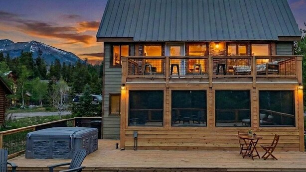 Eine zweistöckige Holzhütte mit einer Terrasse, großen Fenstern und einem Blick auf die Berge.