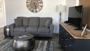 A living room with a grey sofa, a round coffee table, a wooden TV stand, a large clock, and a lamp.