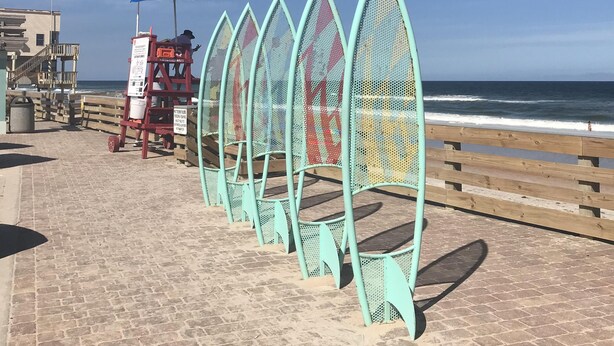 A coastal walkway with a series of green, wave-shaped barriers, a lifeguard stand, and a wooden fence.