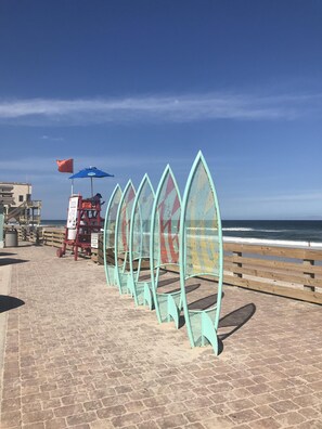 A coastal walkway with a series of green, wave-shaped barriers, a lifeguard stand, and a wooden fence.