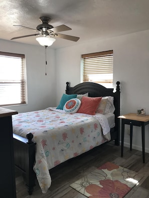 Un dormitorio con cama, ventilador de techo, ventana con persianas, una pequeña mesa de madera y un tapete.