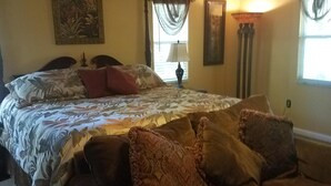 Um quarto com cama, ventilador de teto, janela com cortinas, abajur e um quadro na parede.