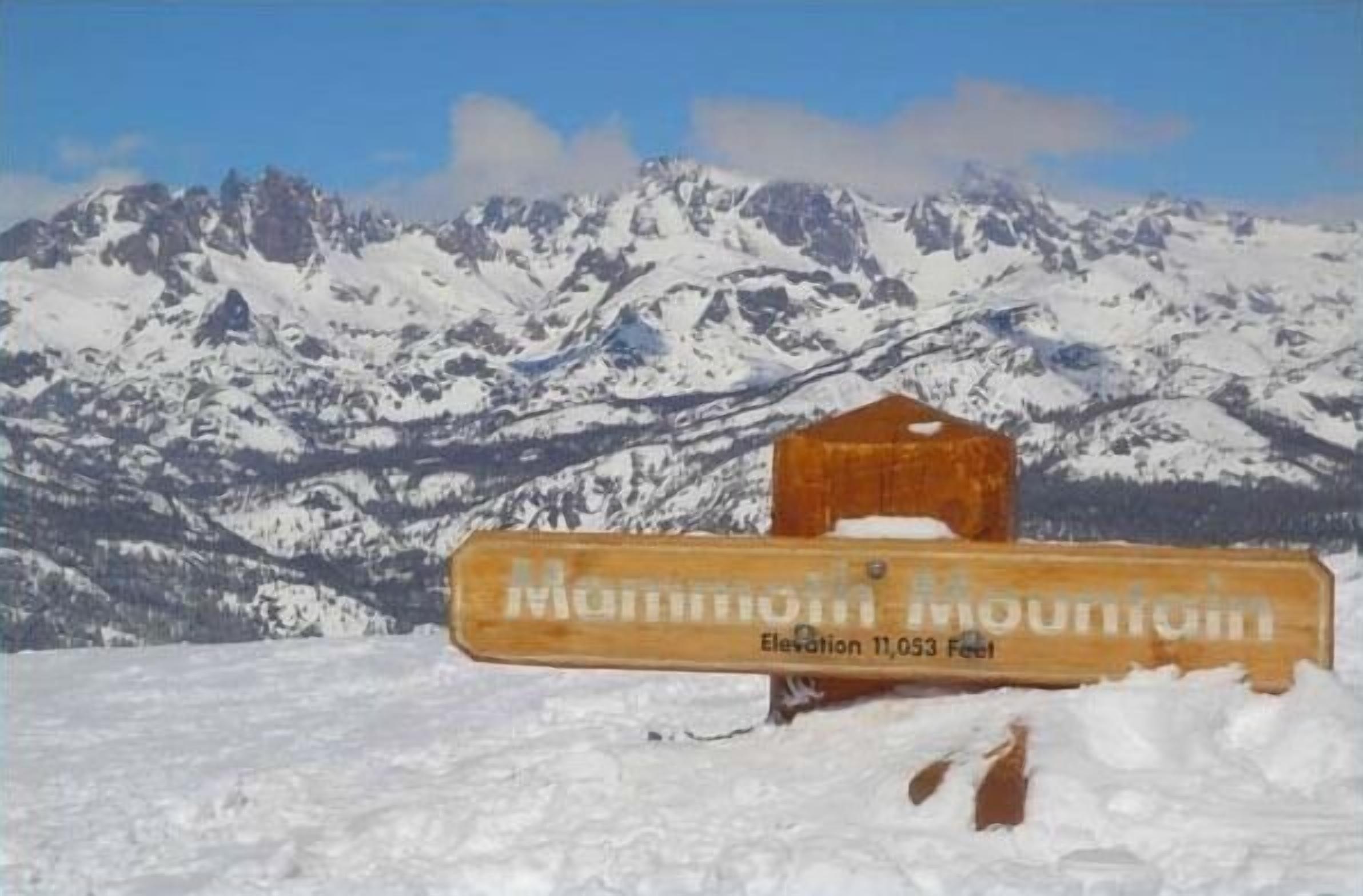 Uma placa de madeira com o texto "Mammoth Mountain" e "Altitude 3.370 metros" ao fundo de montanhas cobertas de neve.