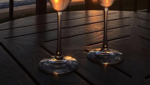 Two glasses of champagne on a wooden table with a view of the ocean and palm trees at sunset. - Remodeled Quiet Oceanfront Corner Unit on 3rd Floor at Sugar Beach Resort (Kihei)