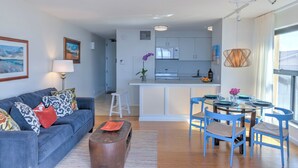 A modern living room with a blue sofa, a coffee table, and a dining area with blue chairs and a glass table. - Remodeled Quiet Oceanfront Corner Unit on 3rd Floor at Sugar Beach Resort (Kihei)