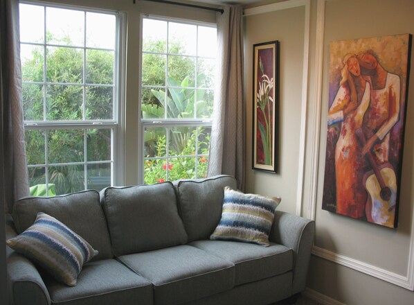 A gray sofa with striped pillows, a framed painting, and a window with a view of greenery.