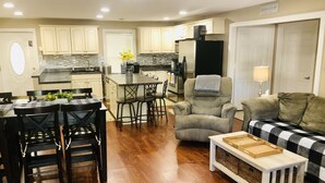 Cozinha moderna com armários brancos, sala de jantar com mesa xadrez preta e branca, e sala de estar com sofá e abajur.