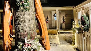 Área externa coberta de neve com uma grande árvore decorada com grinalda e flores, um cercado de madeira e uma casa com entrada iluminada.