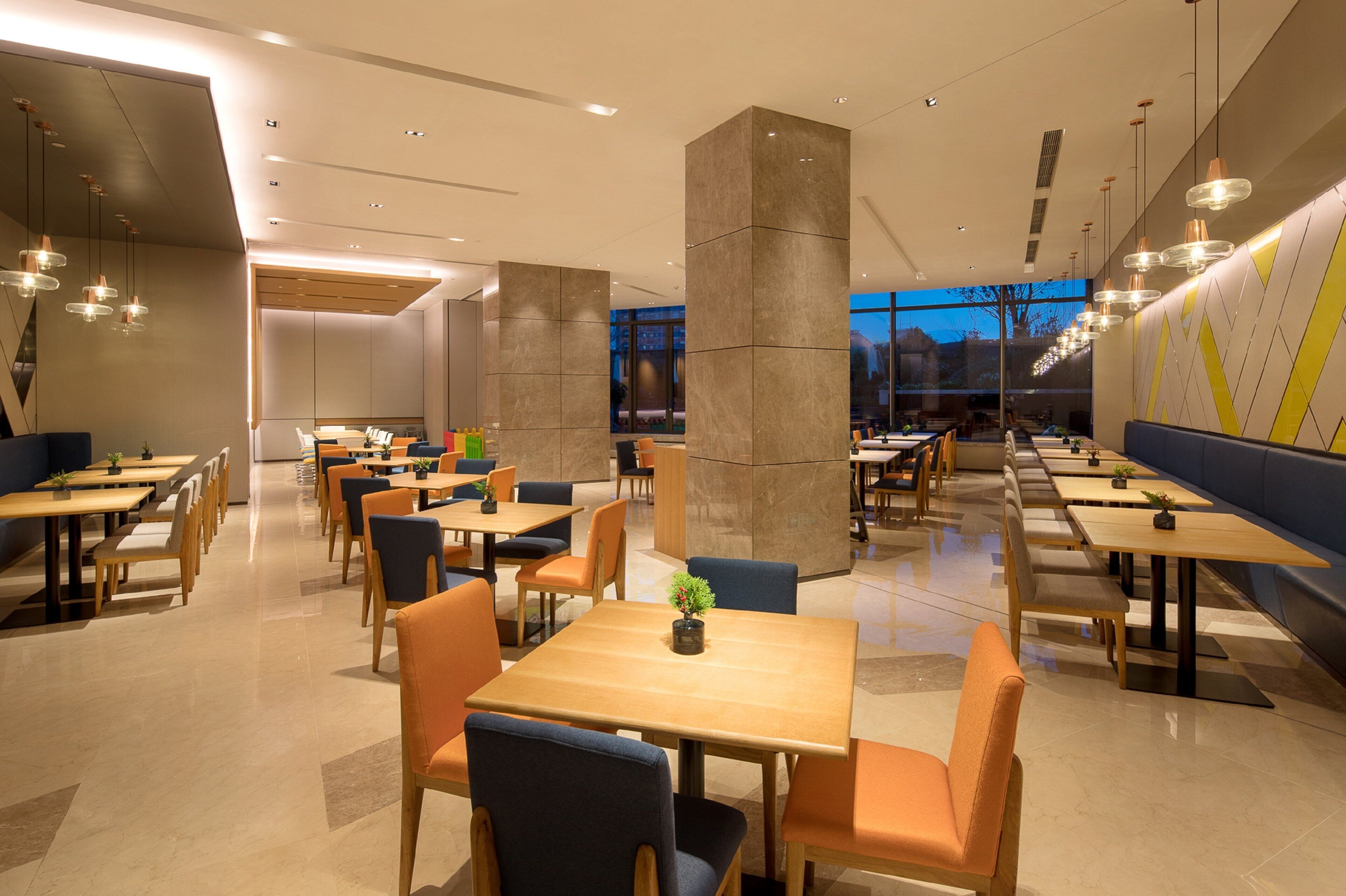 A modern restaurant with multiple tables and chairs, large pillars, and pendant lighting.
