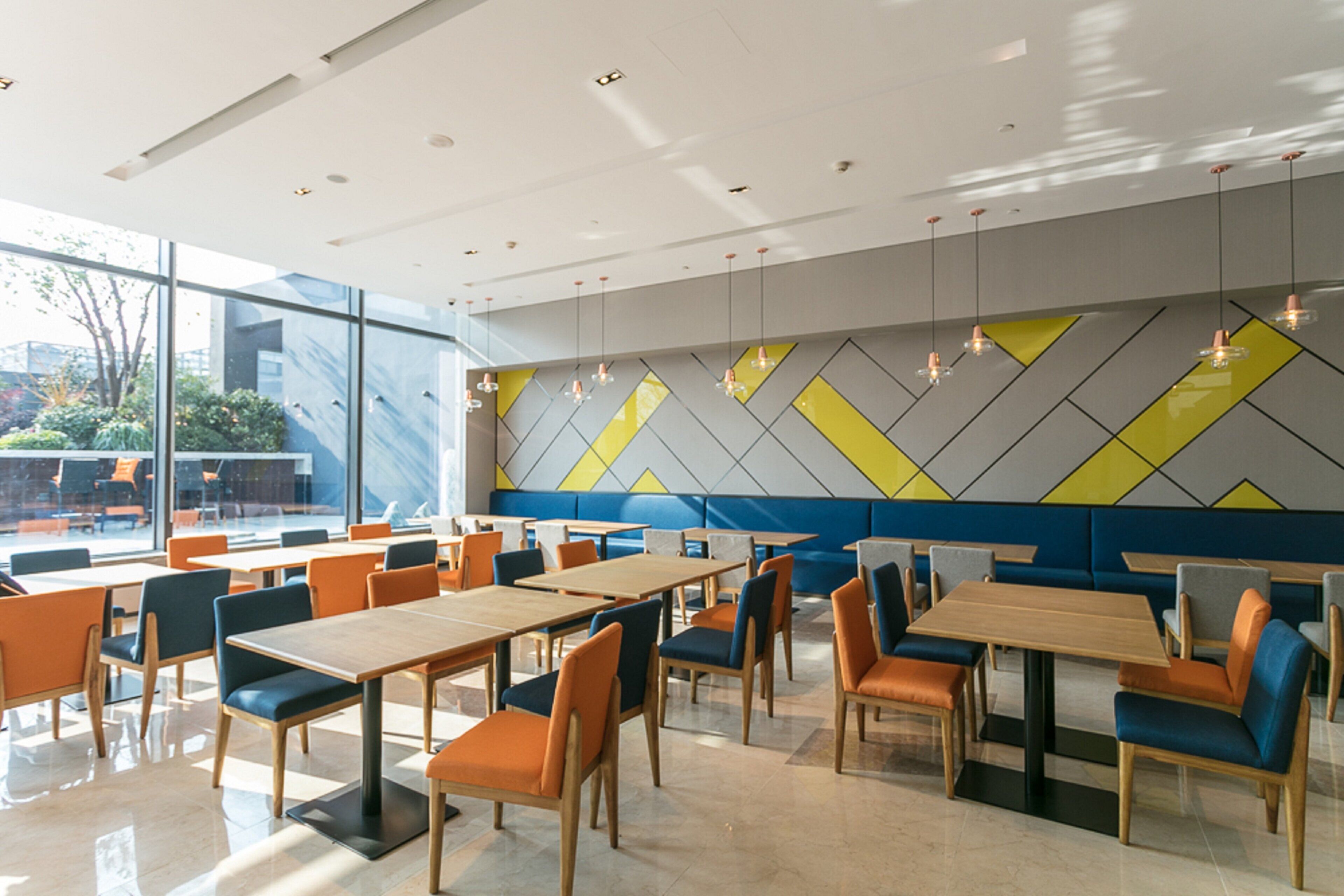 A modern dining area with tables and chairs, large windows, and a geometric wall design.