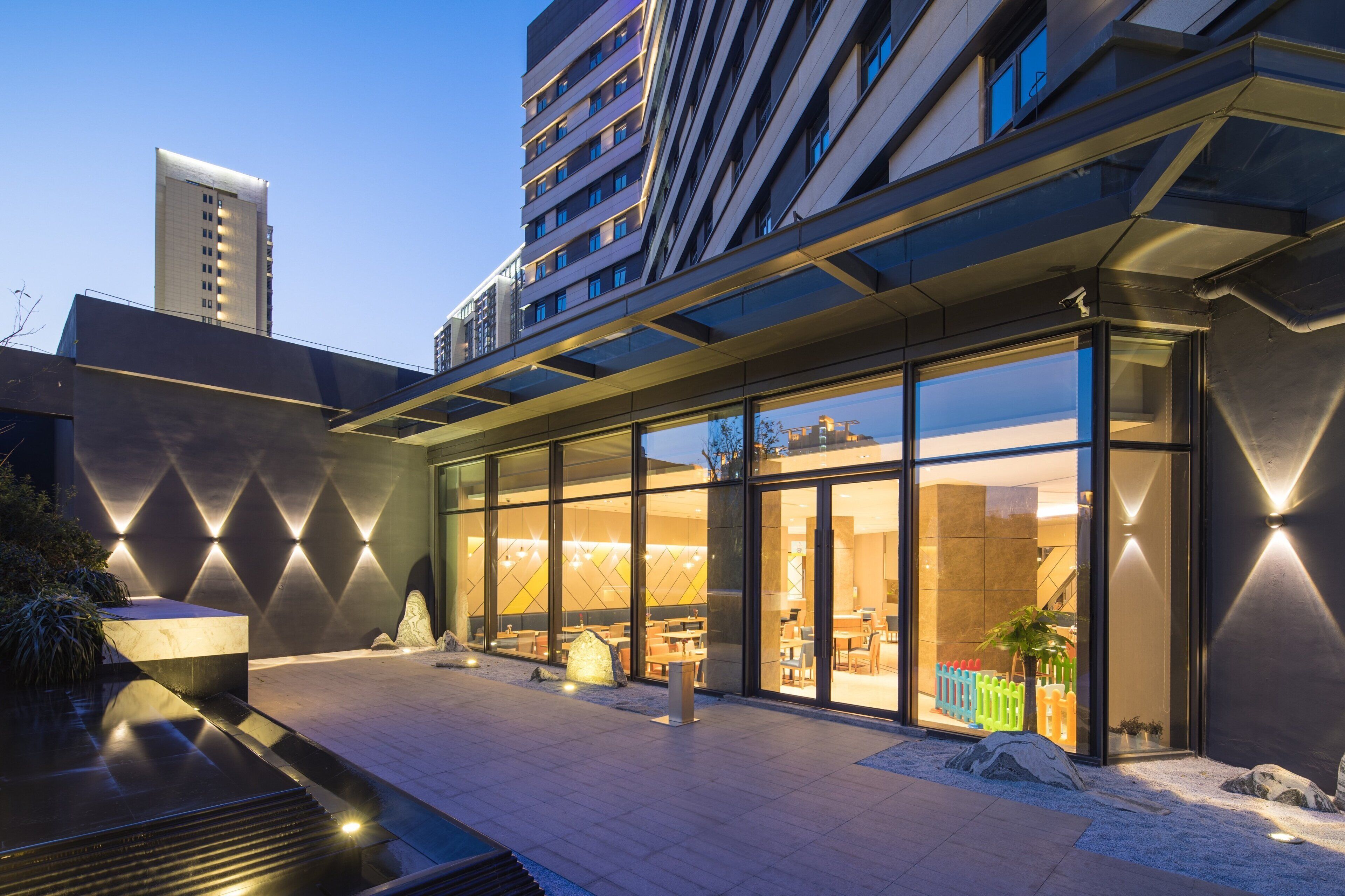 A modern hotel exterior with a glass facade, outdoor seating area, and a landscaped garden.