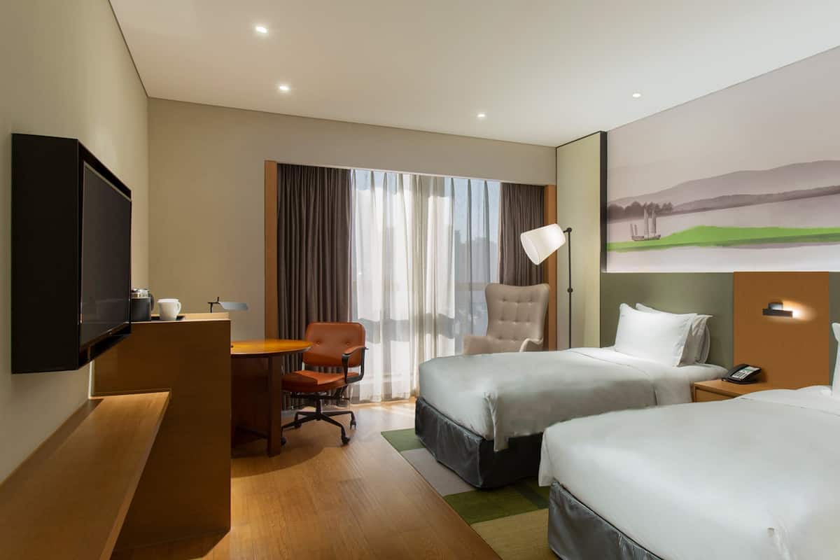 A hotel room with two beds, a desk with a chair, a TV, and a painting of a landscape.