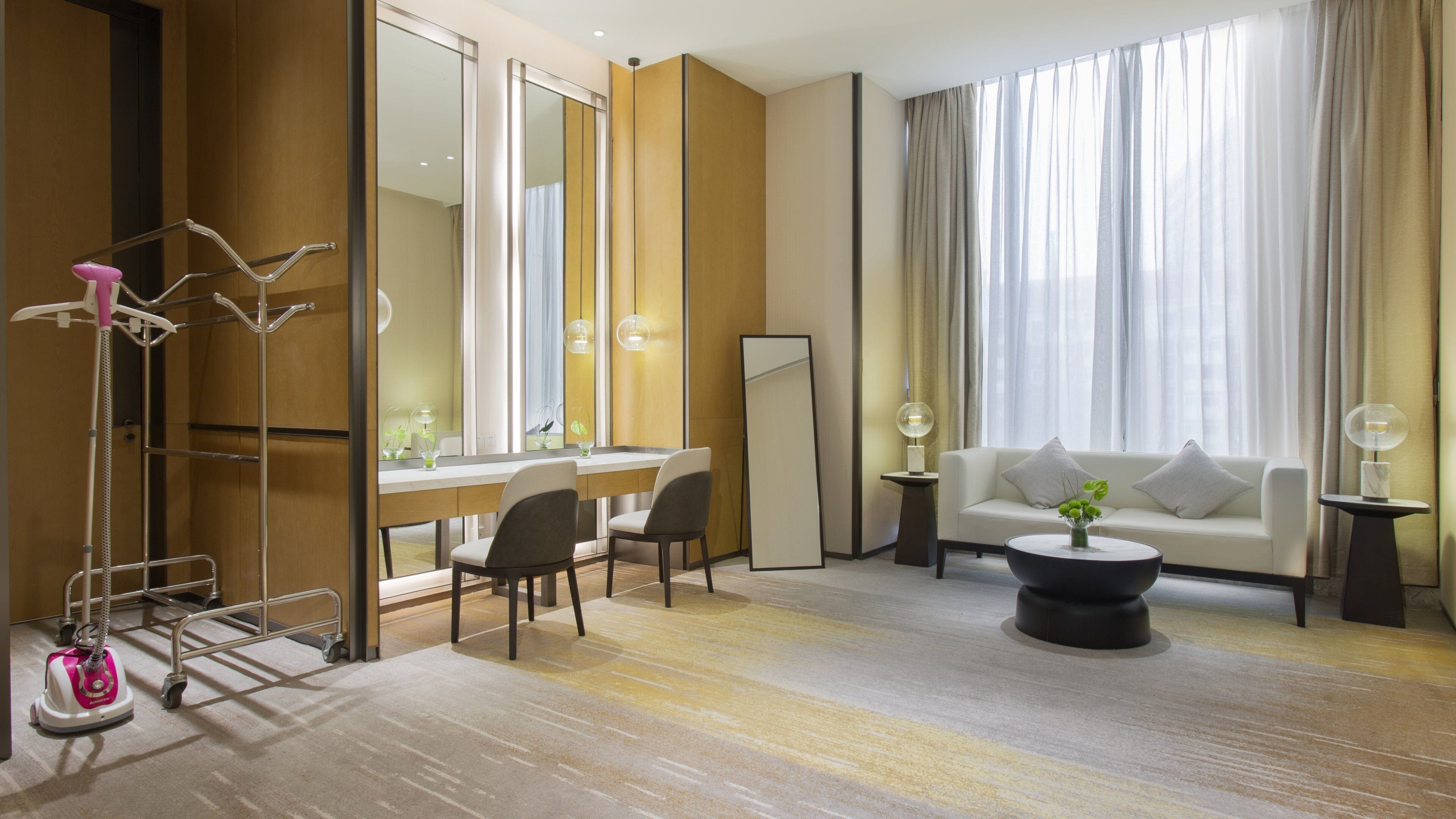 A hotel room with a sofa, a coffee table, a desk with chairs, a large mirror, and a standing heater.
