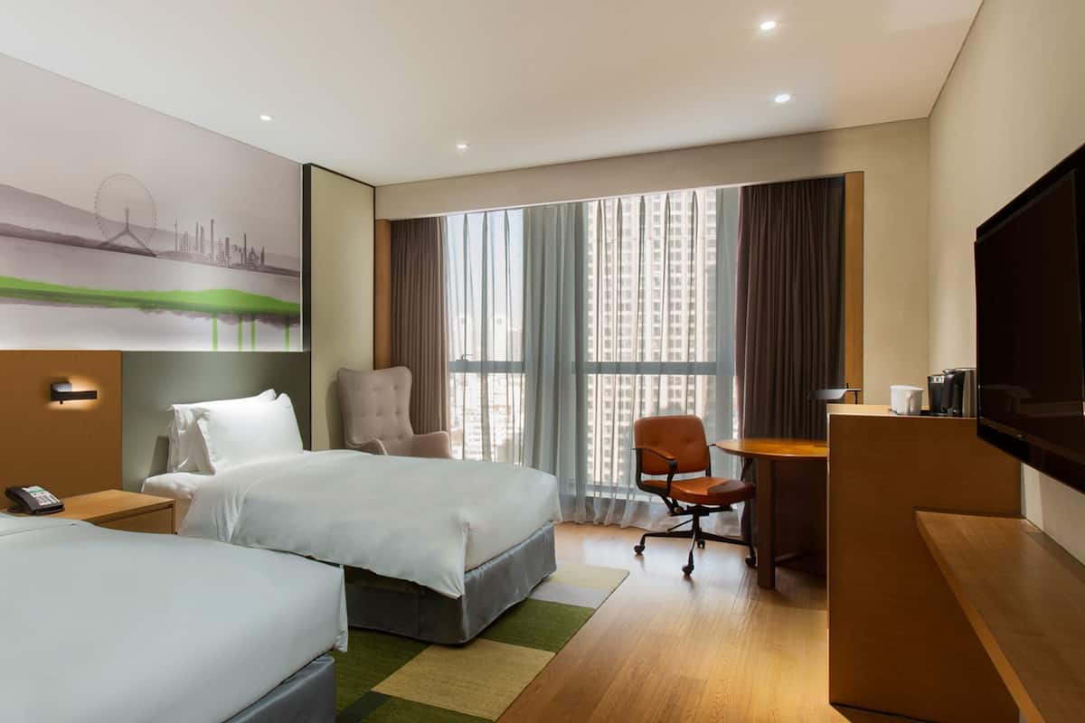 A hotel room with two beds, a desk, a chair, a TV, and a large window with curtains.
