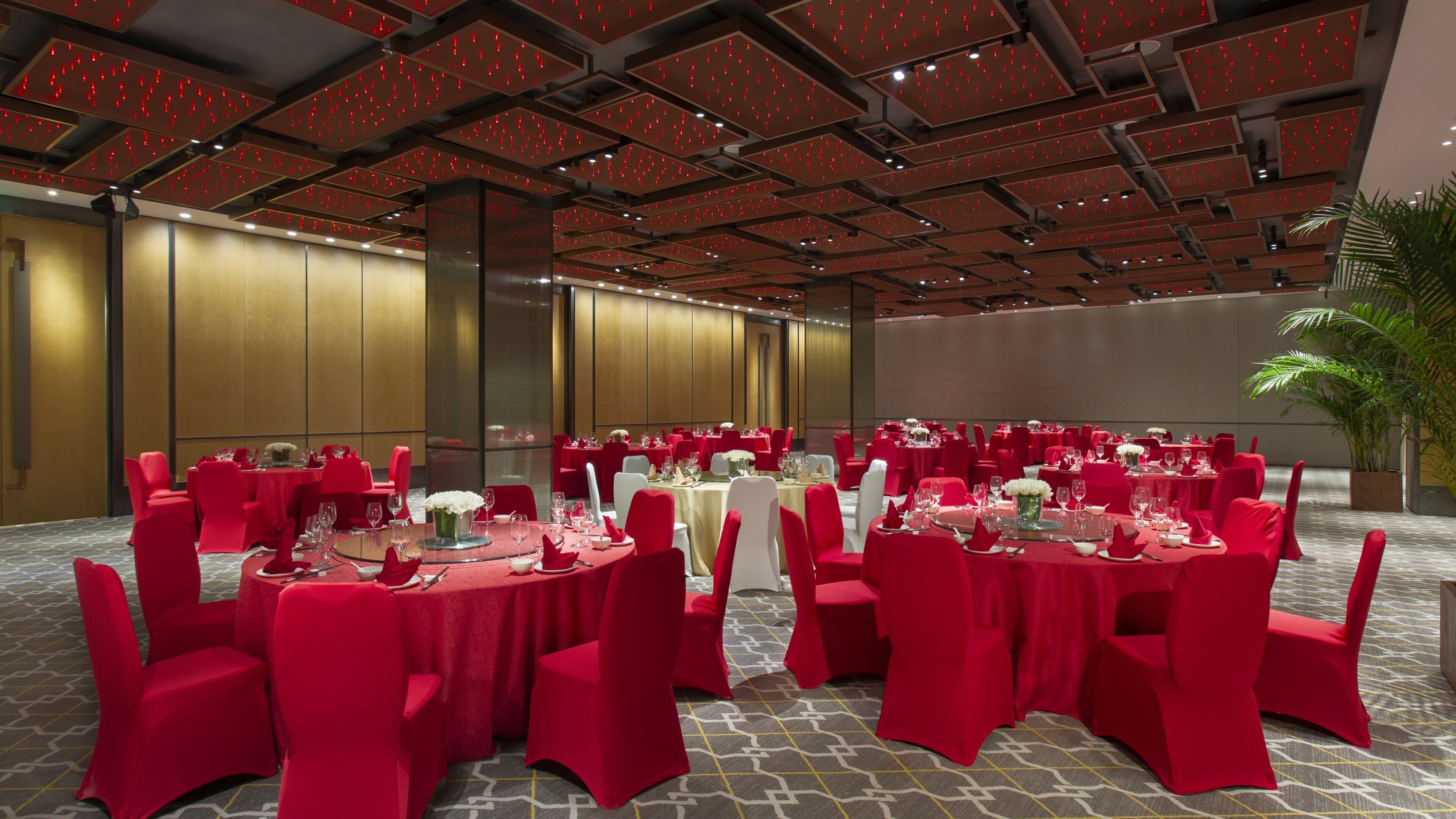 A banquet hall with round tables set for dining, red chairs, and a modern ceiling design.