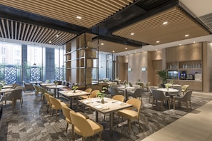 Un espace repas moderne doté de plusieurs tables et chaises, de grandes fenêtres et d’un coin cuisine à l’arrière-plan.