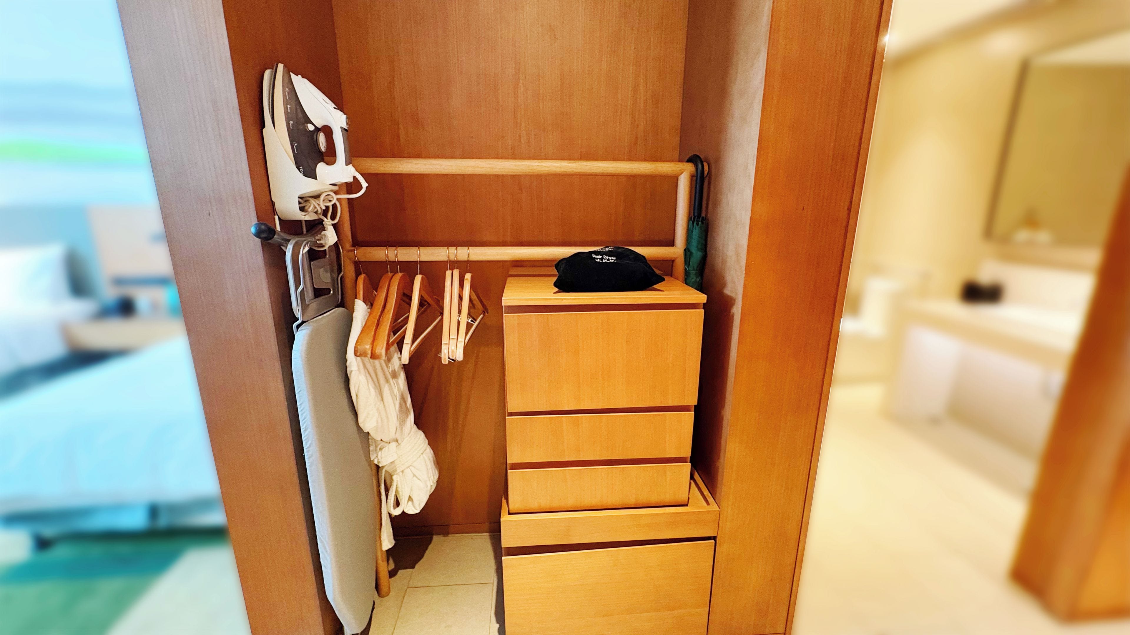 A compact room with a wooden wardrobe, a hanging rack with clothes, and a small table with a cap.