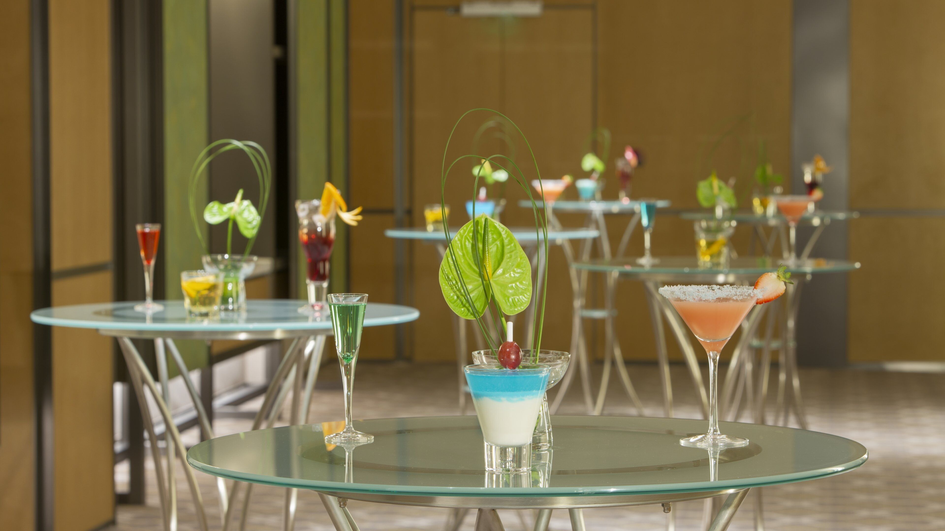A bar area with multiple round glass tables, each with a cocktail and a decorative plant.