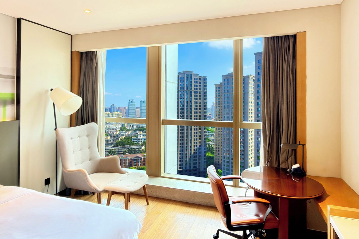 Habitación de hotel con cama, escritorio, silla, lámpara y ventana grande con vista a la ciudad.