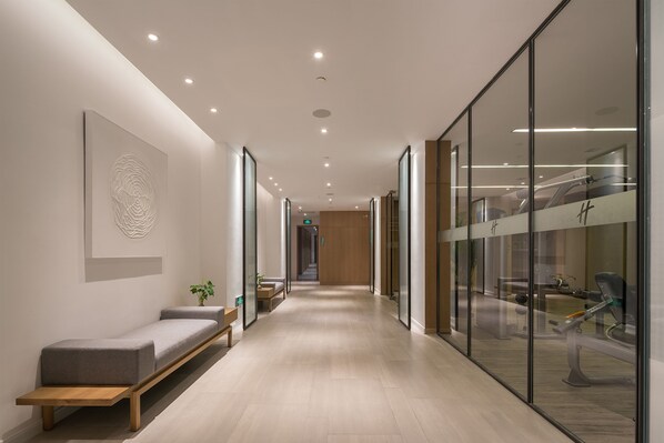 A modern hallway with a bench, a wall-mounted artwork, and a glass wall reflecting gym equipment.