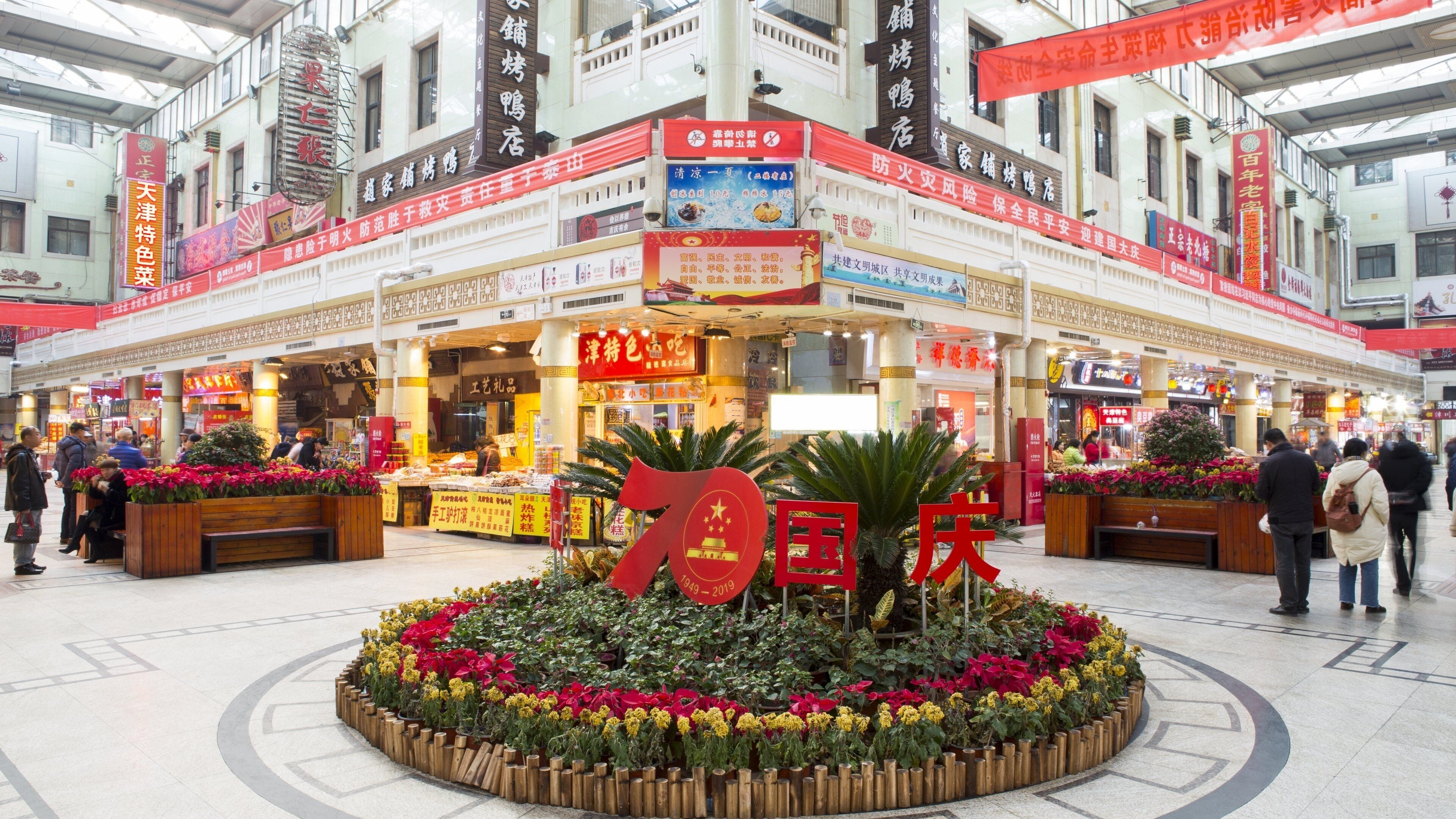 A shopping mall with multiple stores, a central flower display, and a prominent red sign.