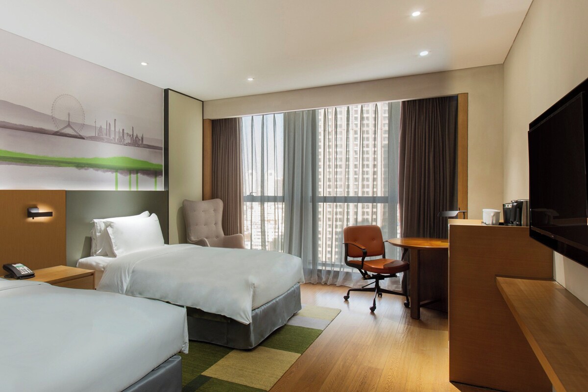 A hotel room with two beds, a desk, a chair, a TV, and a large window with curtains.