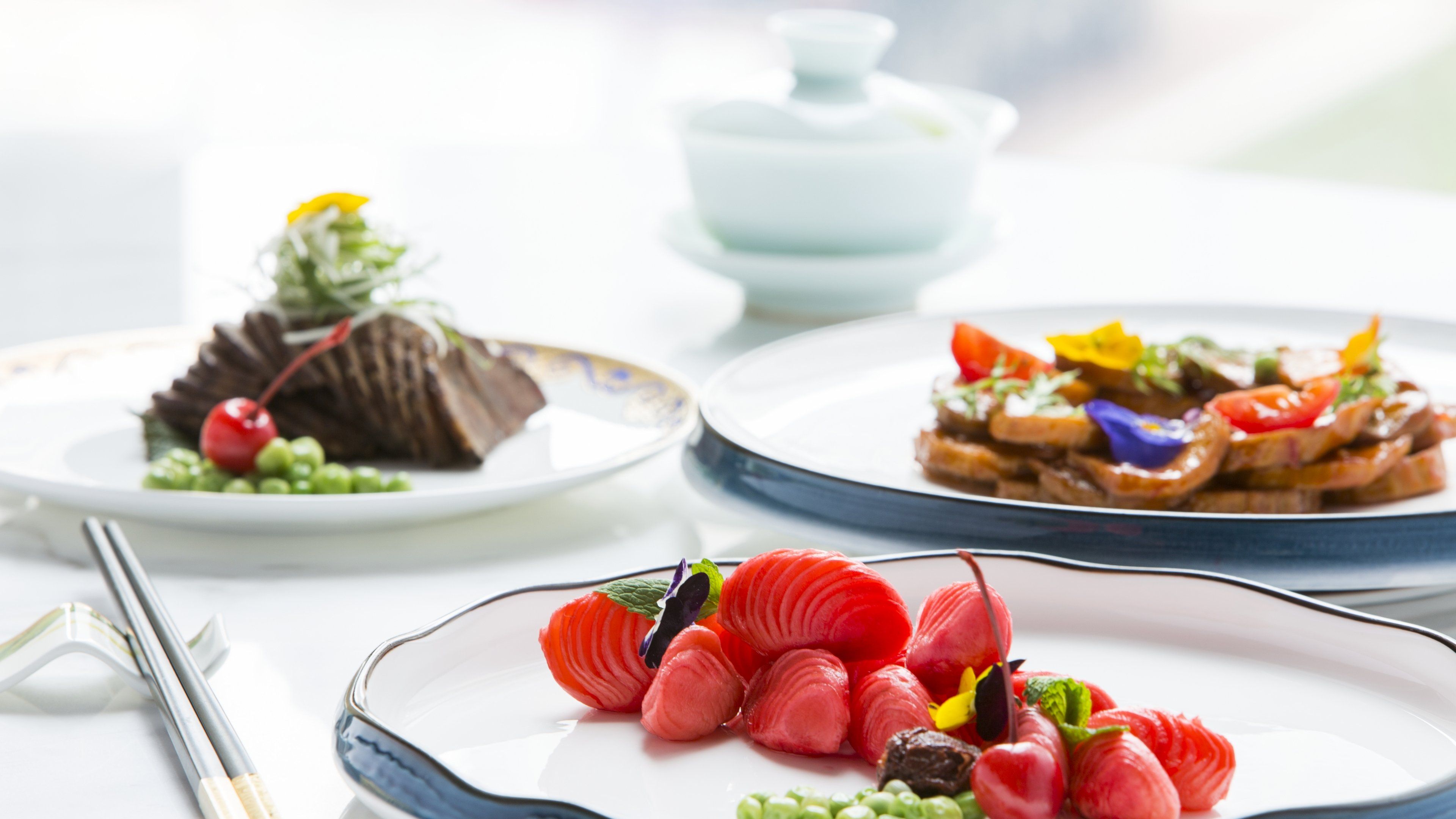 Three plates of food with chopsticks and a teapot on a table.