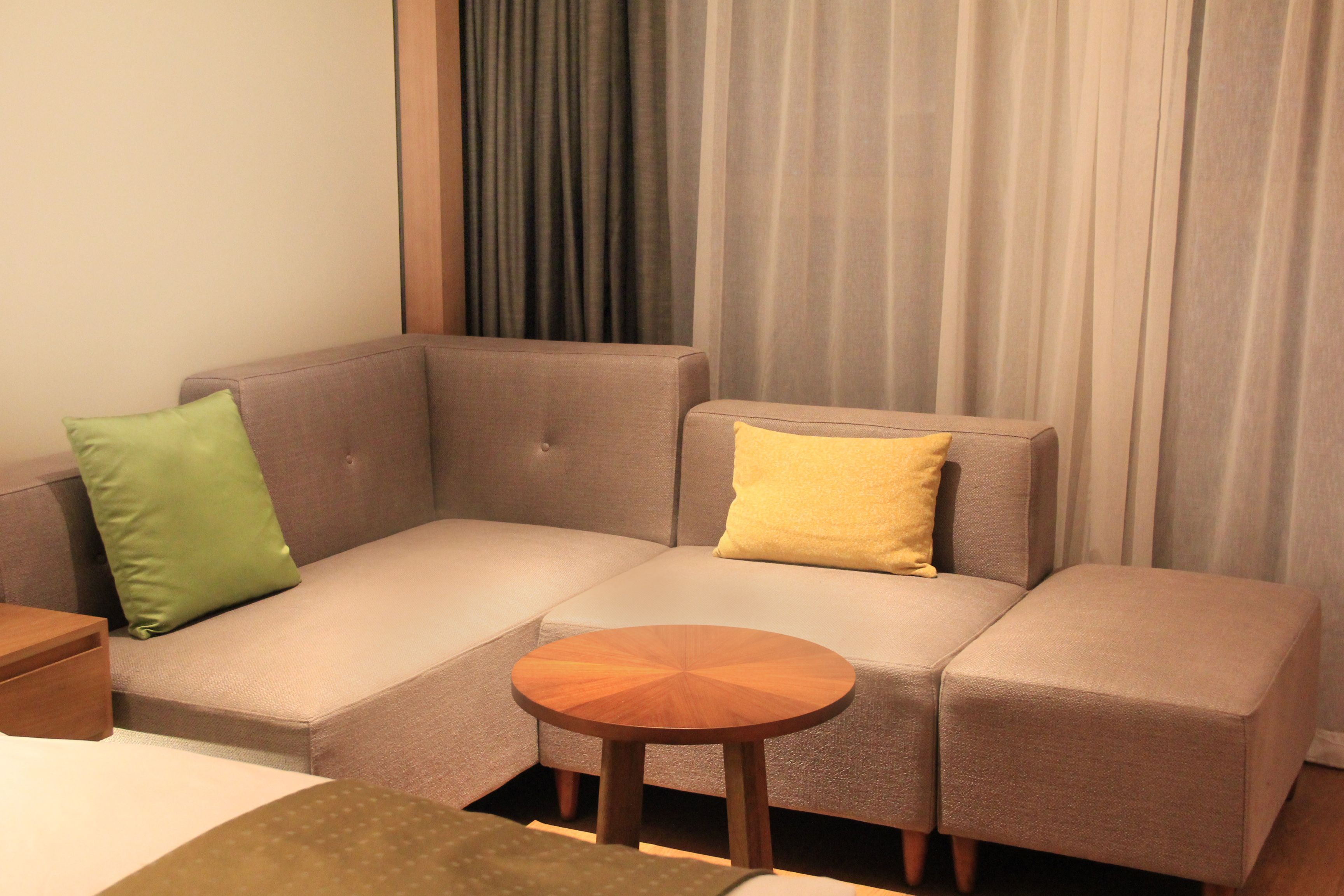 A corner sofa with a small wooden side table, a green and a yellow pillow, and a window with curtains.