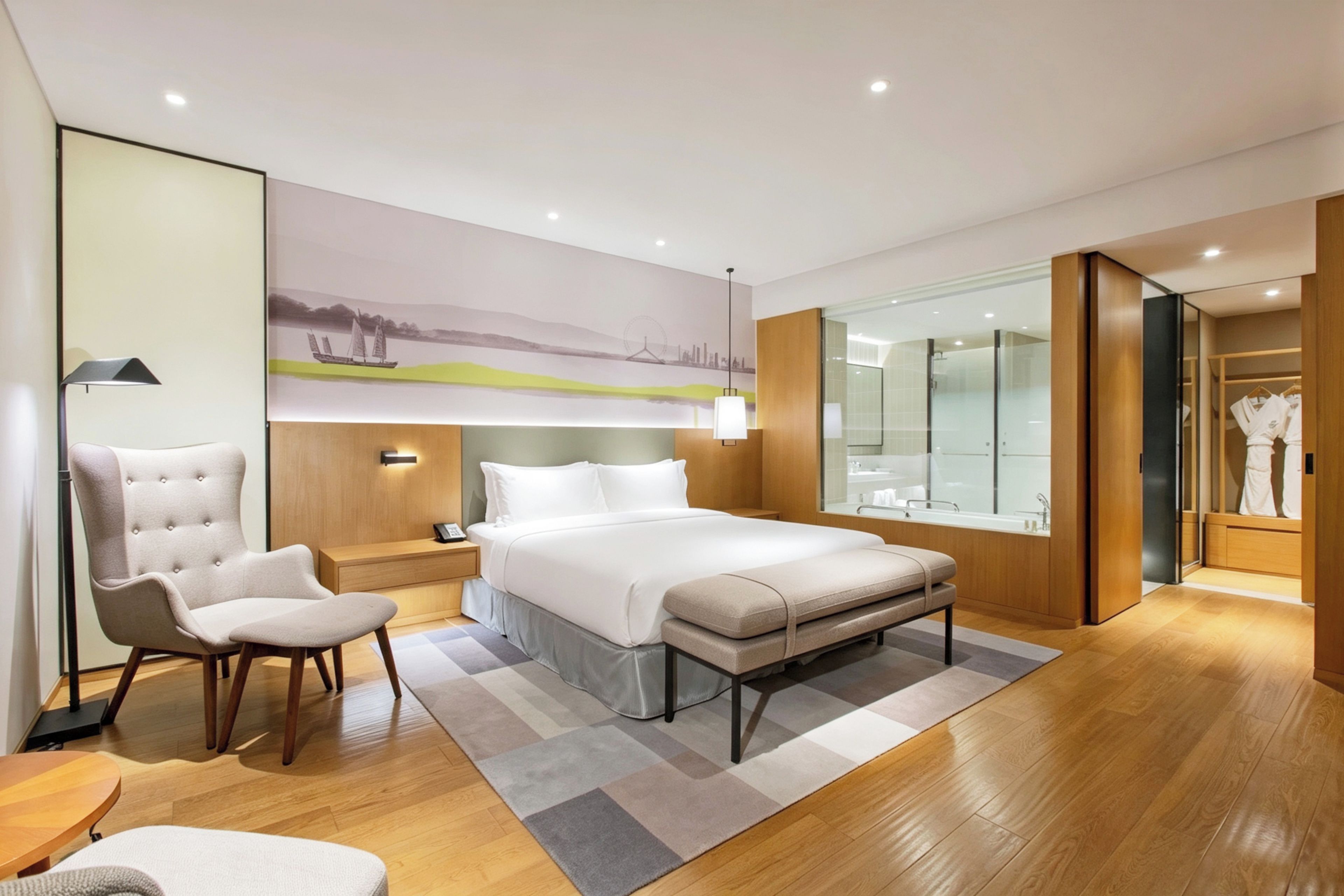 Una habitación de hotel moderna con una cama grande, una zona de estar con una silla, una mesita de noche y una lámpara de pared, además de un espejo amplio.