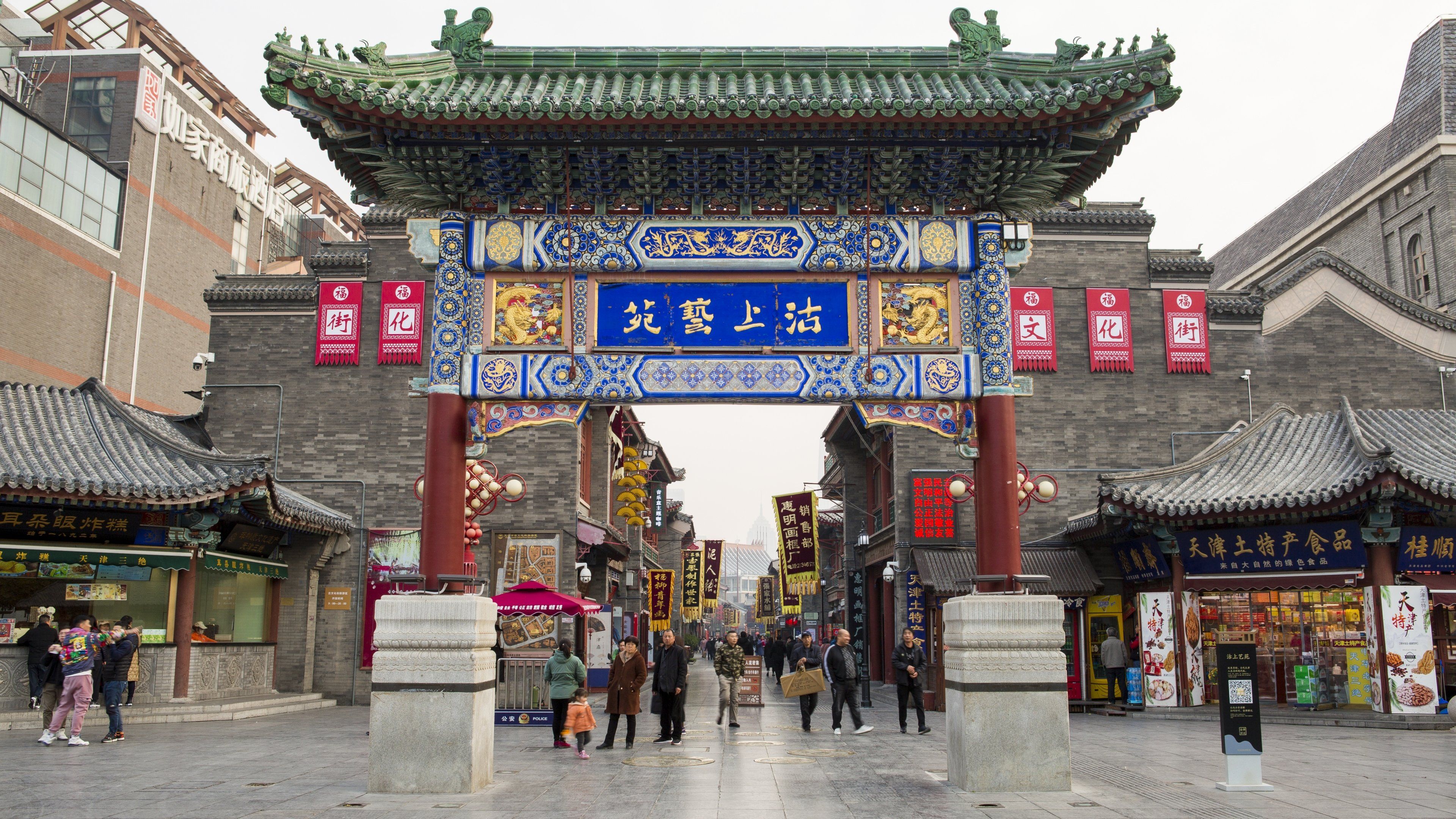 Una puerta tradicional china con diseños intrincados en azul y dorado, que da paso a una calle bordeada de tiendas y restaurantes.