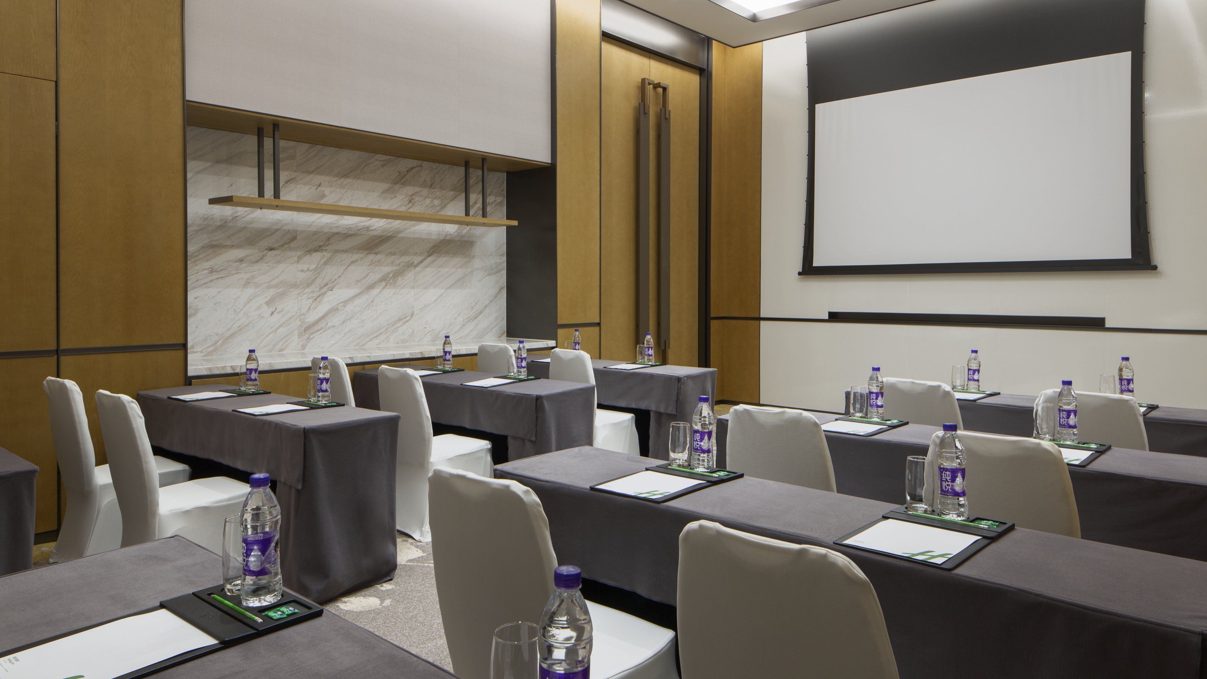 Un salón de conferencias con mesas, sillas, botellas de agua y una pantalla de proyector.