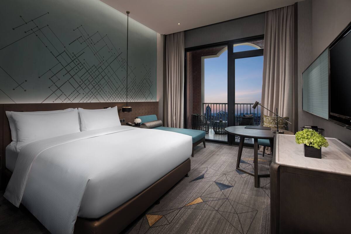 Een moderne hotelkamer met een groot bed, een bureau, een stoel, een bijzettafeltje, een kamerplant en een groot raam met uitzicht.