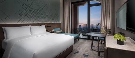 Una habitación de hotel moderna con una cama grande, un escritorio, una silla, una mesita pequeña, una planta en maceta y un ventanal con vistas.