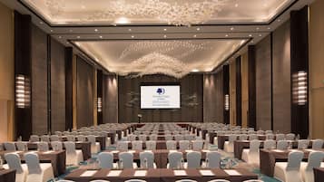 Un salón de conferencias con filas de mesas y sillas, una pantalla grande al frente y un techo con una lámpara decorativa.
