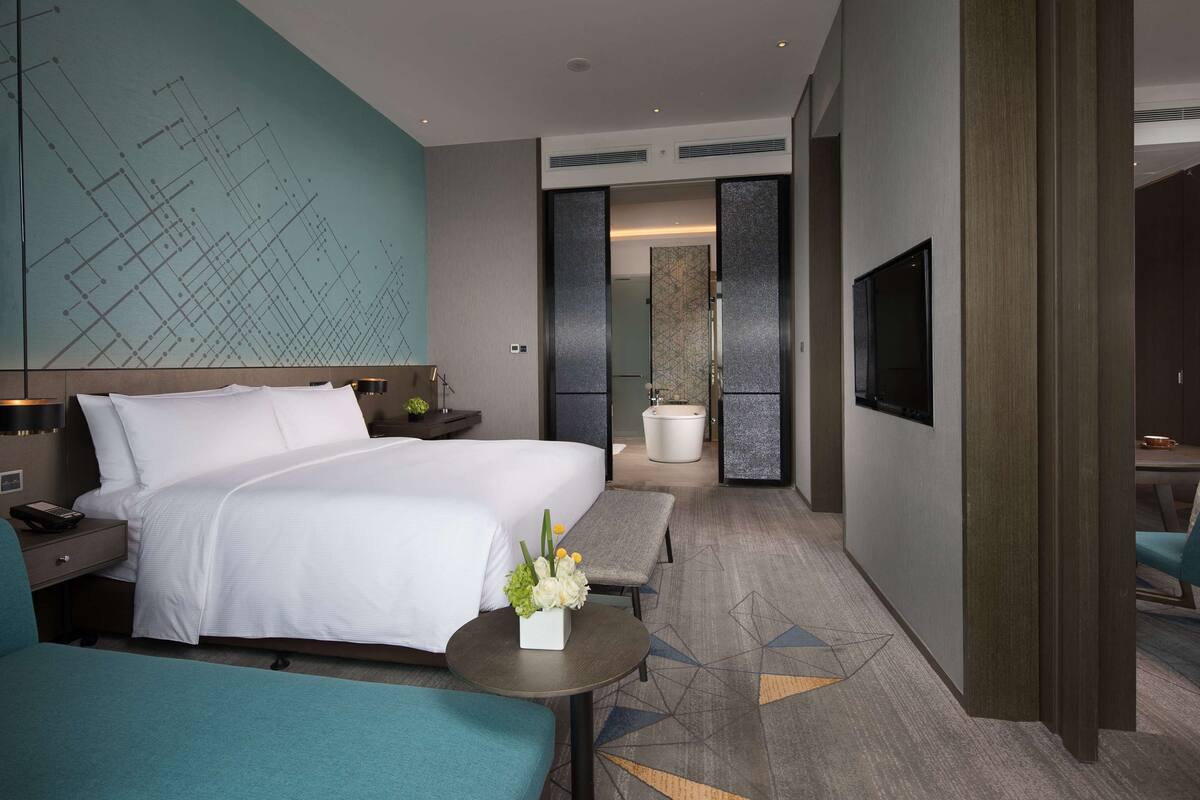 Een moderne hotelkamer met een groot bed, een bureau, een televisie en een badkamer die zichtbaar is door een glazen deur.