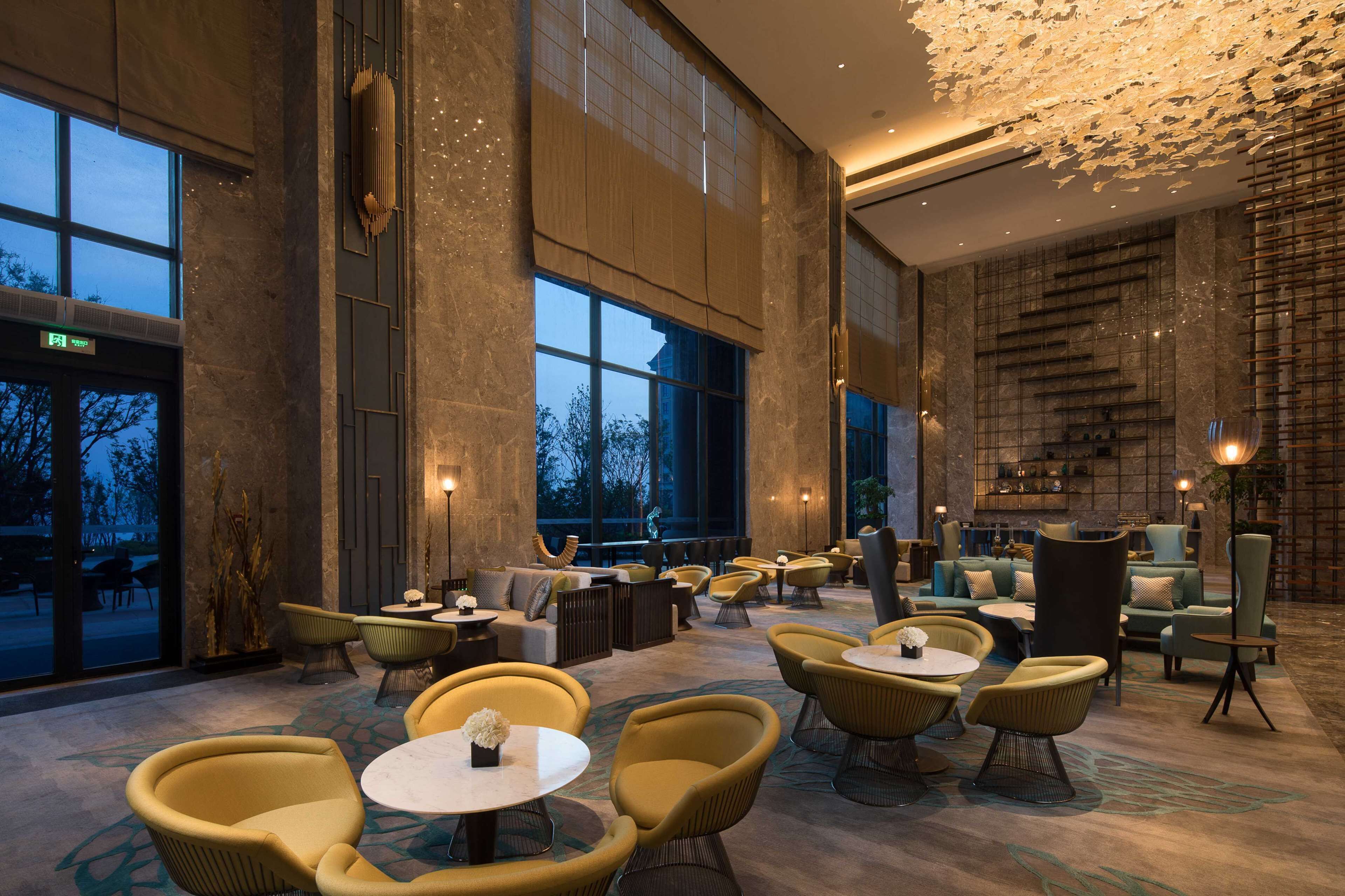 Een ruime lobby met hoge plafonds, grote ramen en diverse zithoeken, waaronder stoelen en banken.