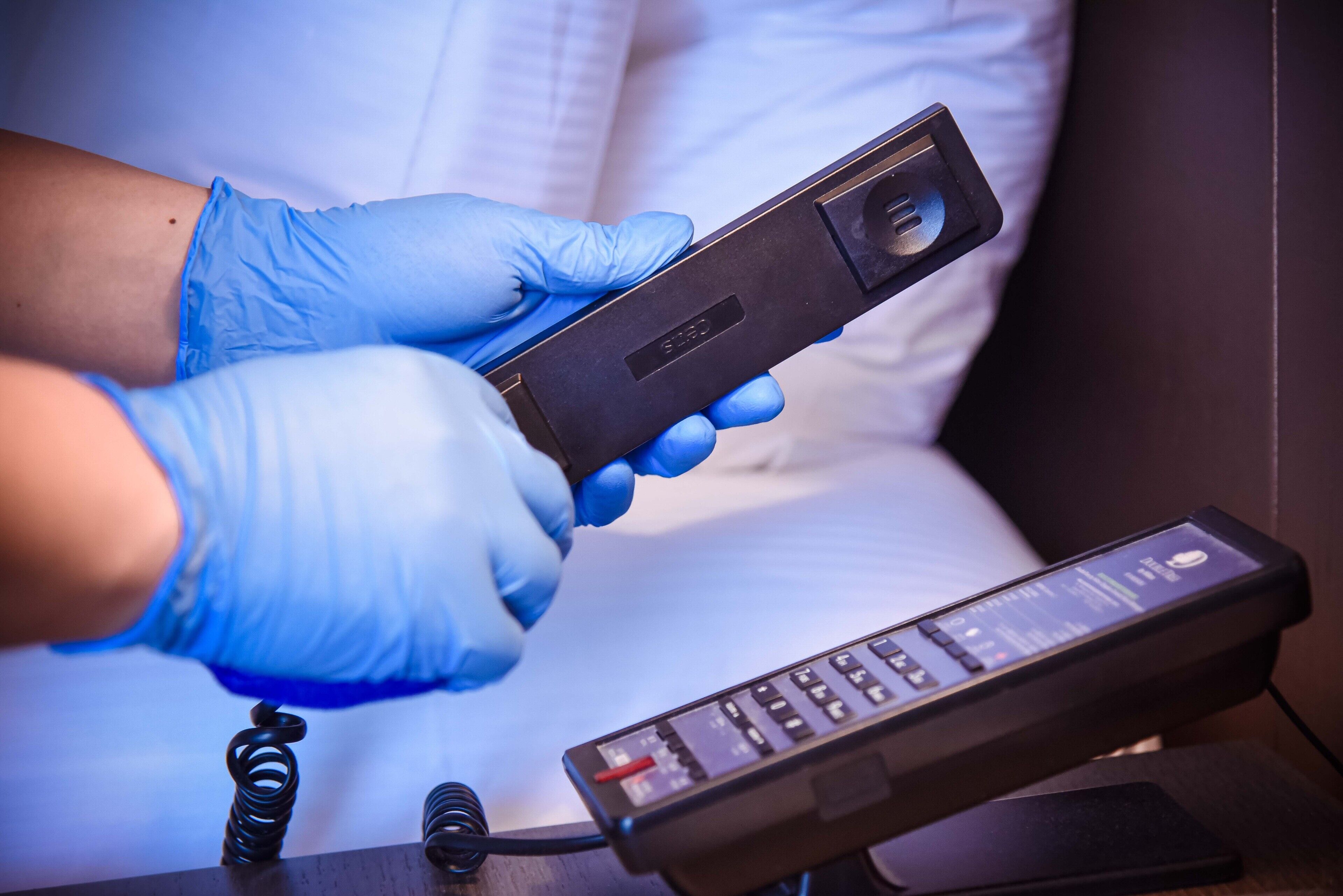 A person in blue gloves holding a black device with a screen and buttons, likely a medical or technical instrument, in a room with a white bed and a black remote control on a table.