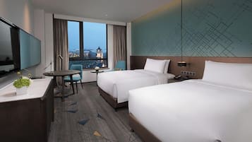 Habitación de hotel con dos camas, un escritorio, una silla, un televisor y un ventanal con vistas a la ciudad.