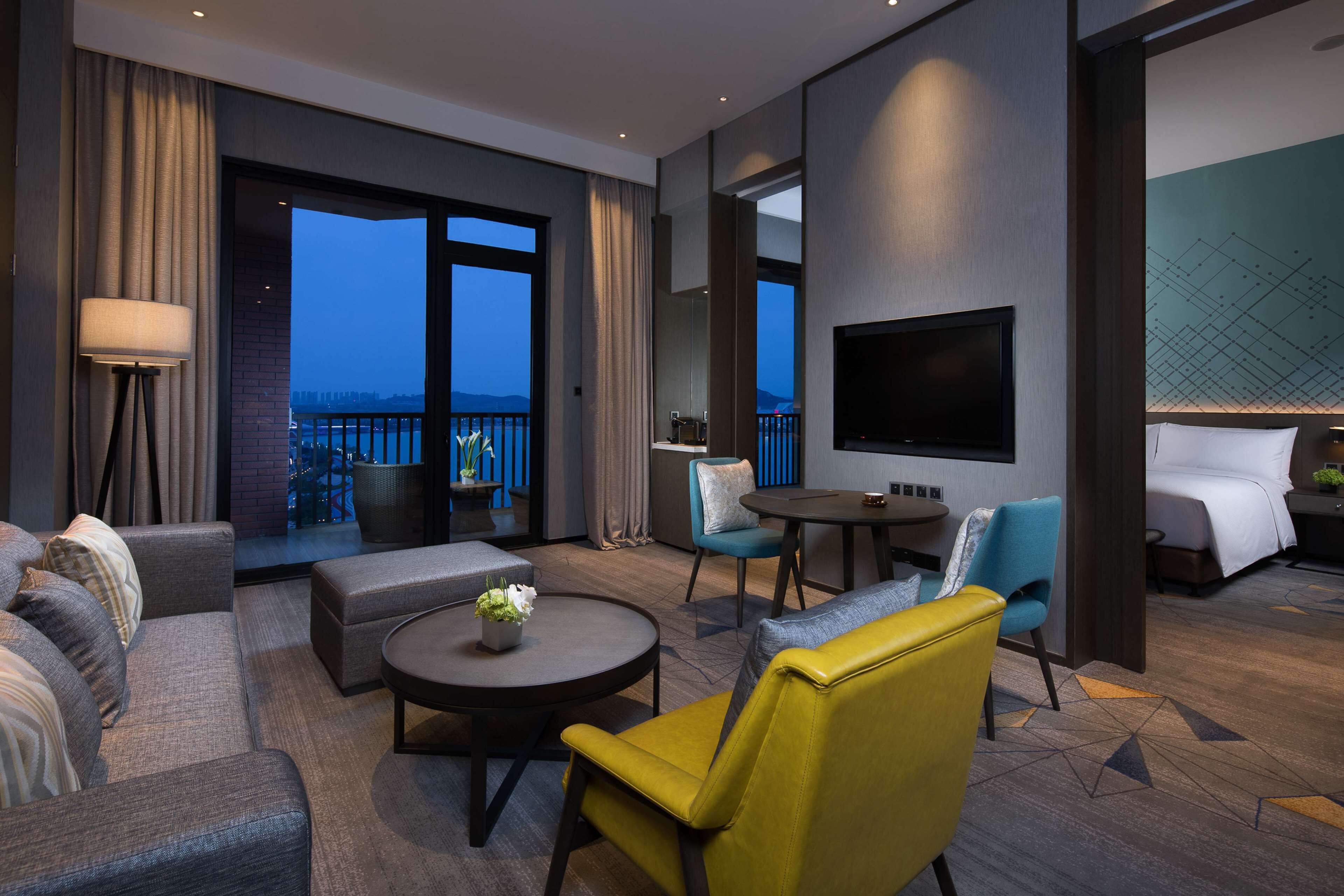 Habitación de hotel moderna con balcón, televisor de pantalla plana, sofá, sillón amarillo y mesa de comedor.