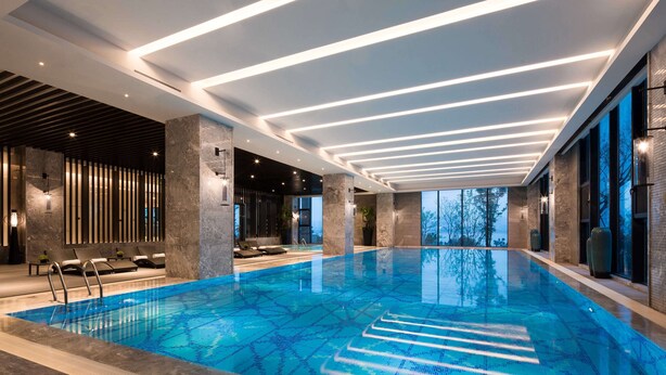 A modern indoor swimming pool with clear water, geometric ceiling lighting, and a view of trees outside. - DoubleTree by Hilton Qingdao Oriental Movie Metropolis (Qingdao)
