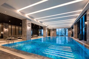 Une piscine intérieure moderne, dotée d’une eau cristalline, d’un éclairage au plafond aux formes géométriques et offrant une vue sur les arbres à l’extérieur.