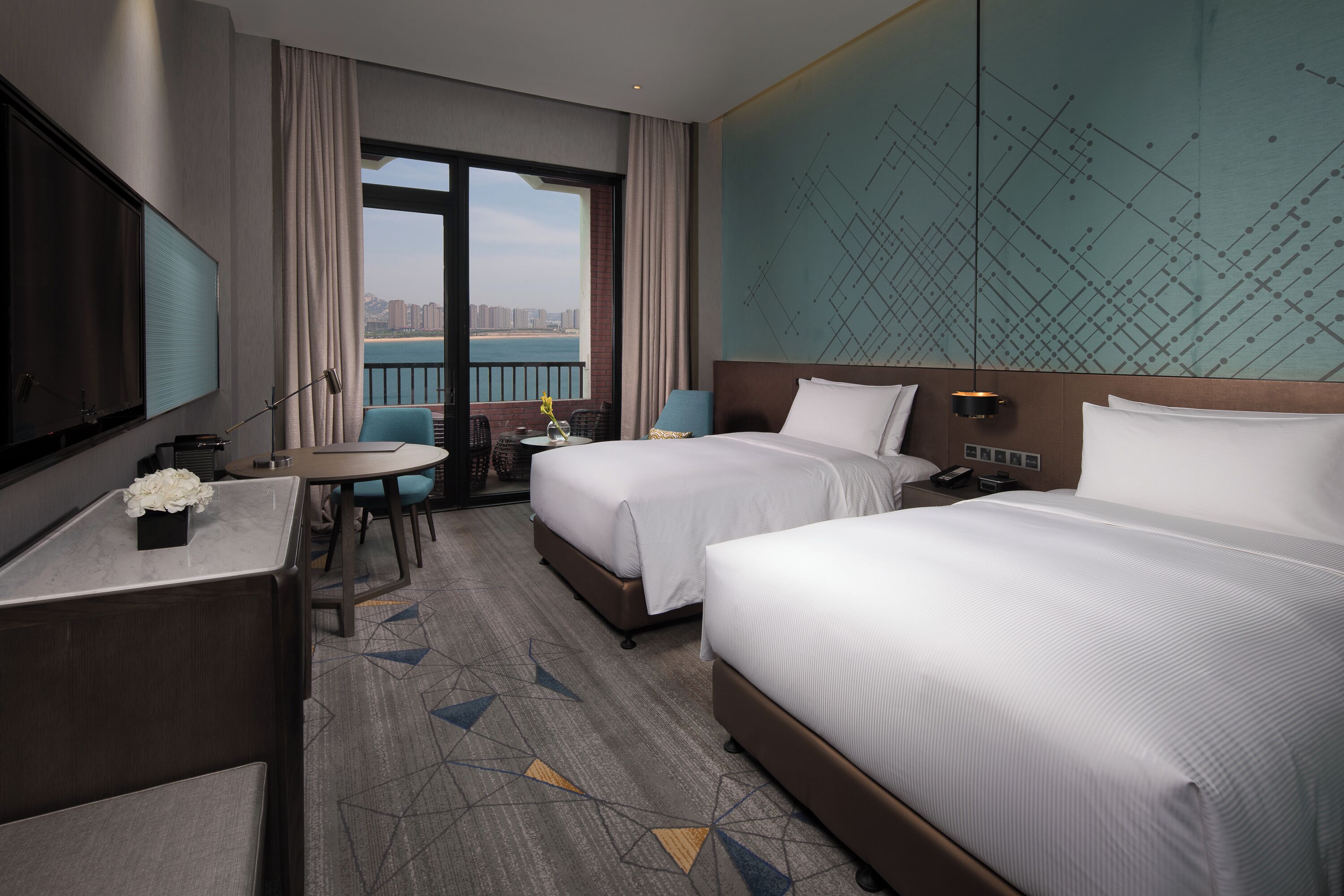 A hotel room with two beds, a desk with a TV, a balcony with a view of the city, and a patterned carpet.