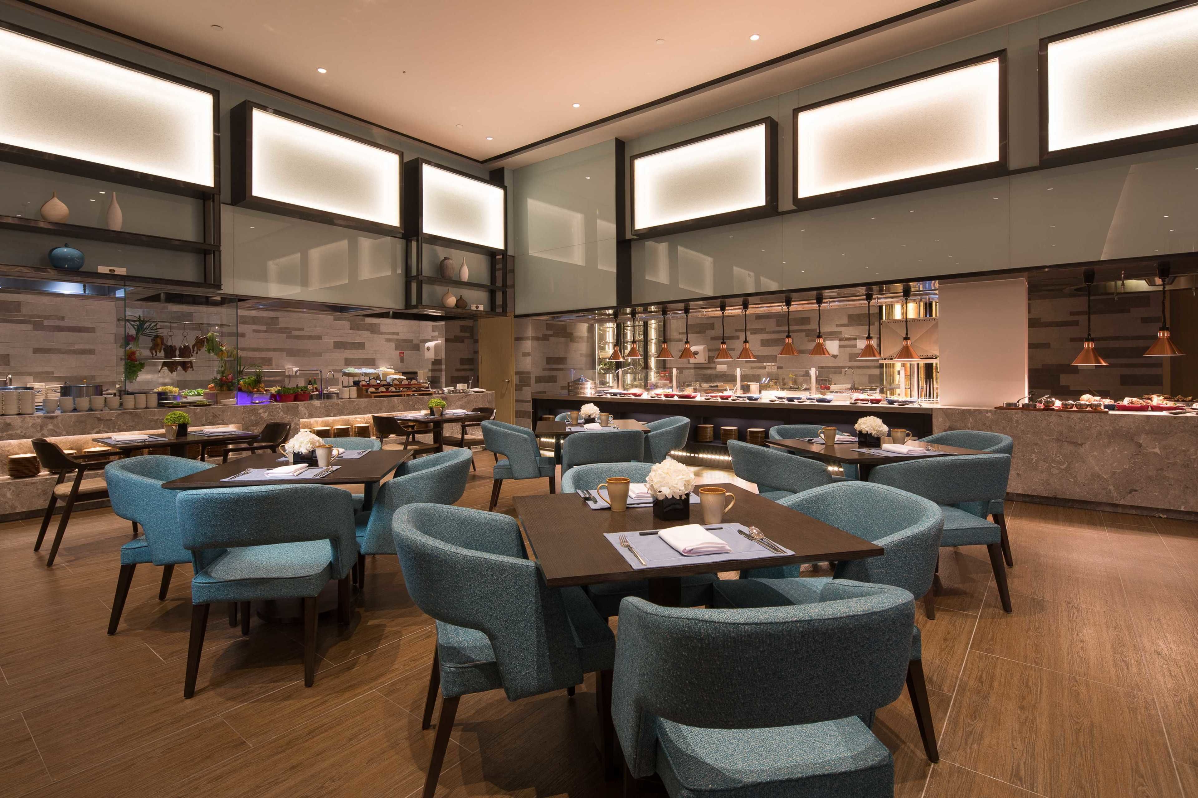 A modern restaurant with teal chairs and dark tables, featuring a kitchen area with copper pendant lights and a bar counter.