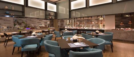 A modern restaurant with teal chairs and dark tables, featuring a kitchen area with copper pendant lights and a bar counter.