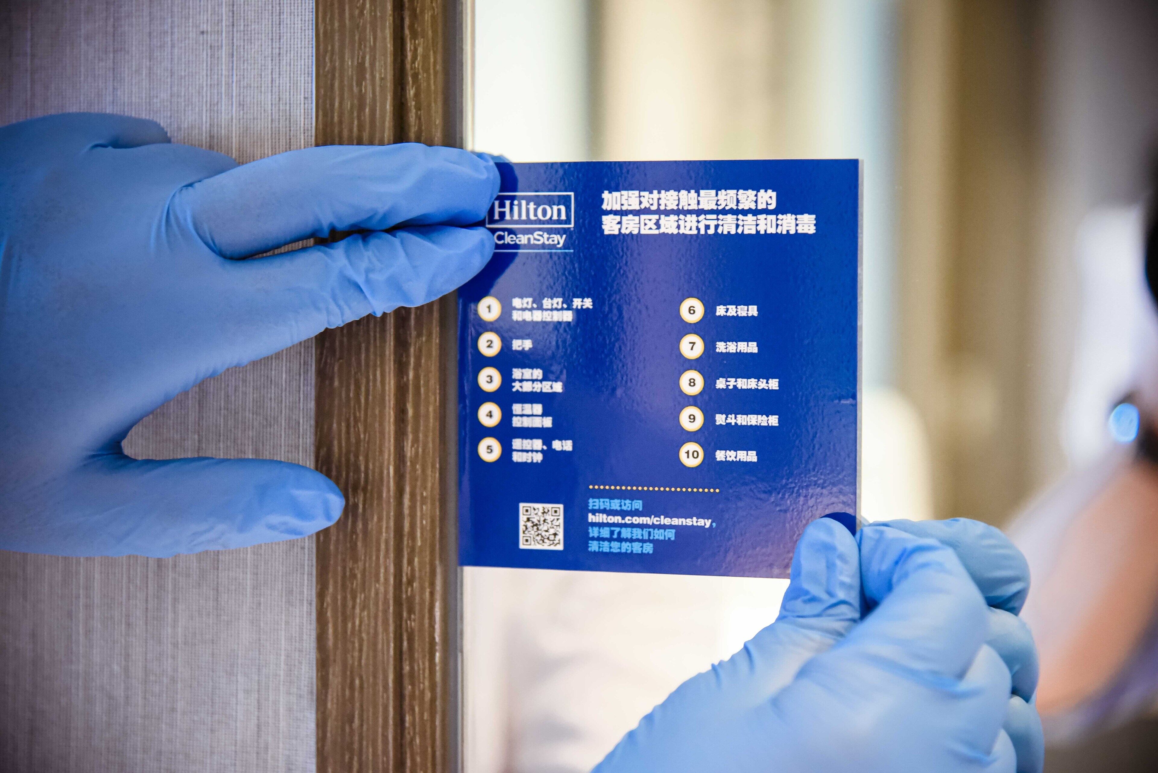 Eine Person mit blauen Handschuhen hält eine blaue Karte mit Text und einem QR-Code, vermutlich im Zusammenhang mit Hotelservices oder -informationen.
