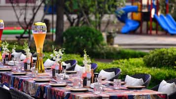 An outdoor dining area with tables set for a meal, surrounded by greenery and a playground in the background.