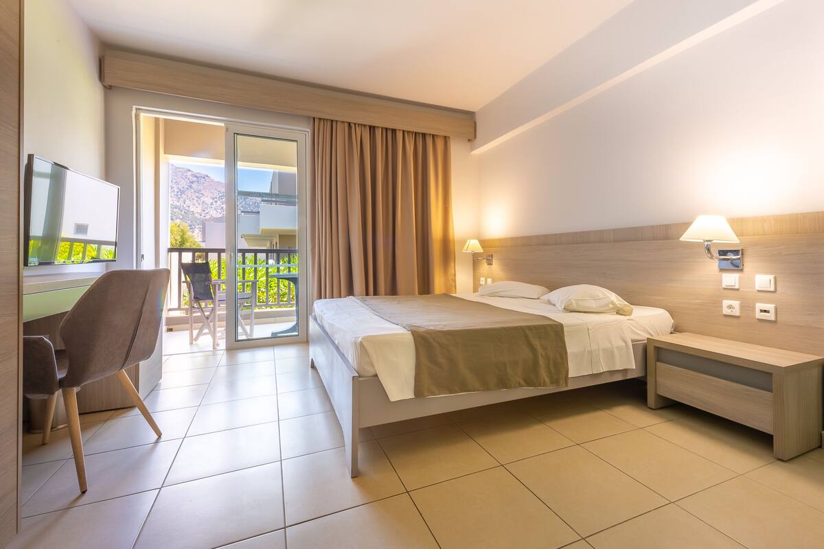 A hotel room with a bed, bedside tables, a chair, a TV, and a balcony with curtains.