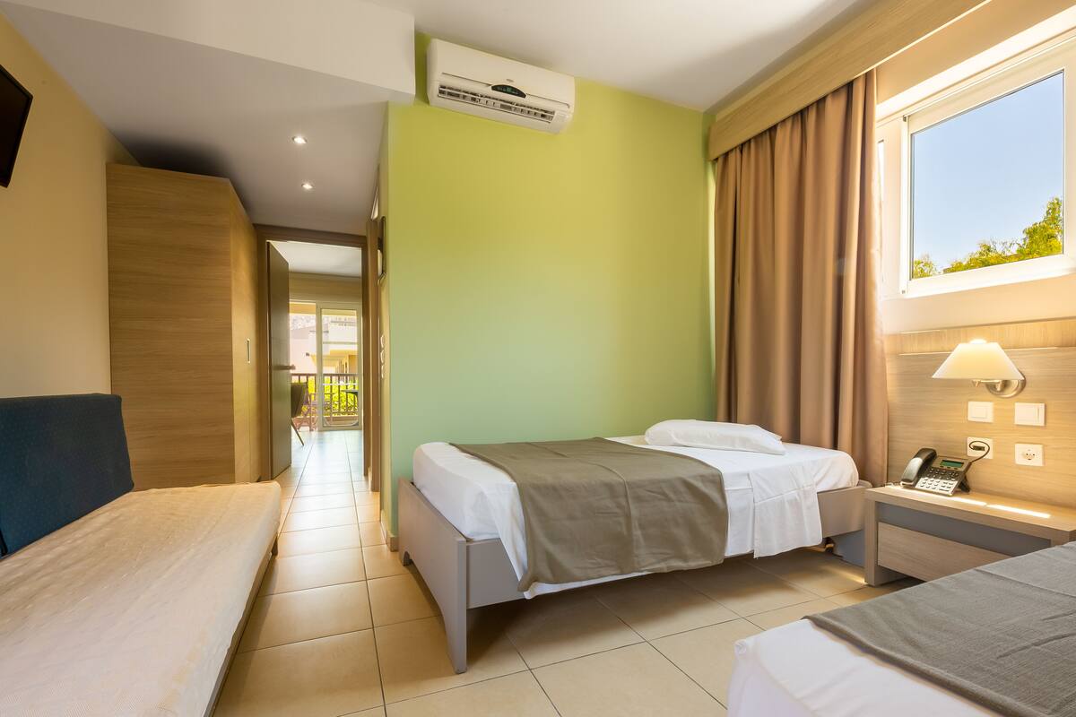 A hotel room with a bed, bedside table with a phone, a window with curtains, an air conditioning unit, and a TV on the wall.
