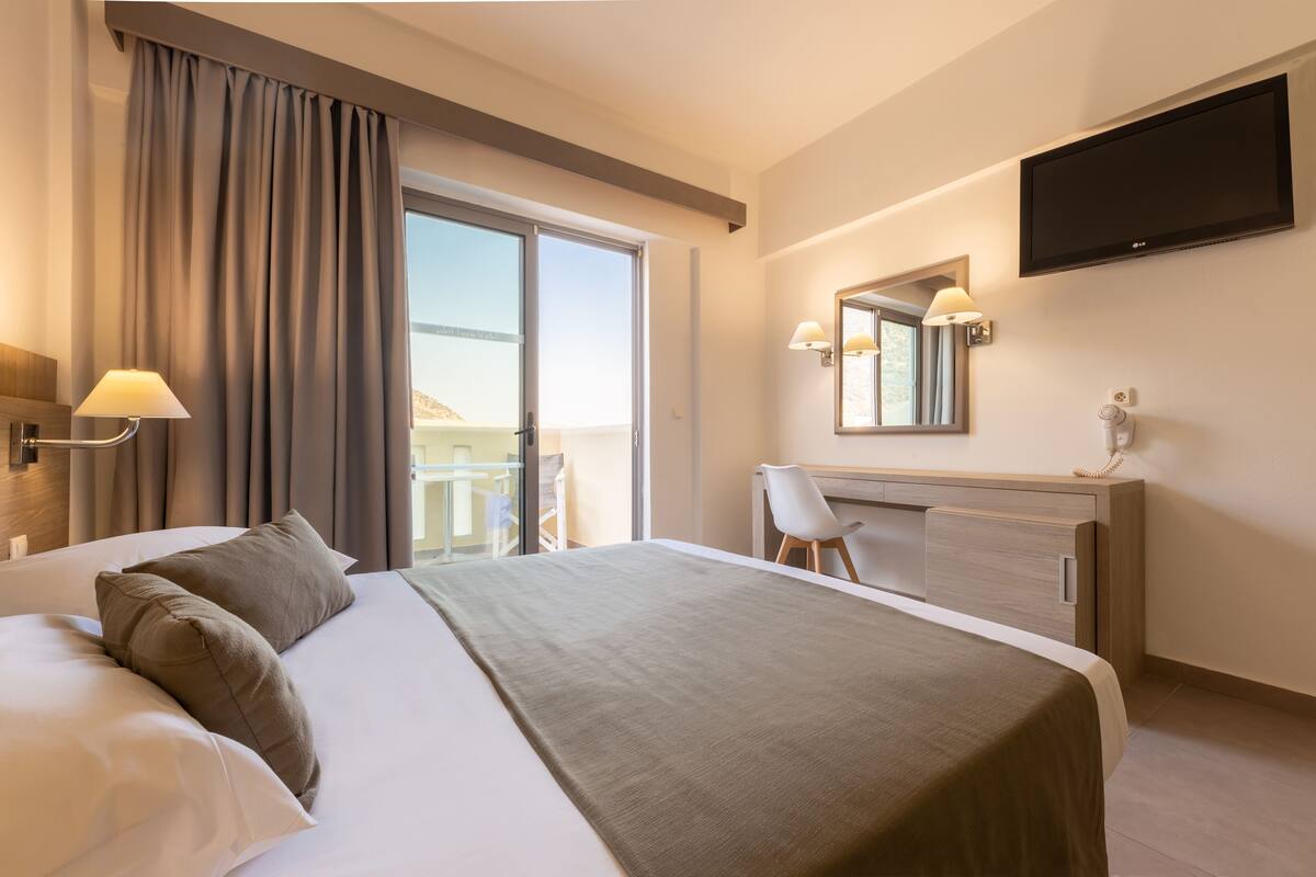 A hotel room with a large bed, a desk, a chair, a TV, and a balcony with a view.