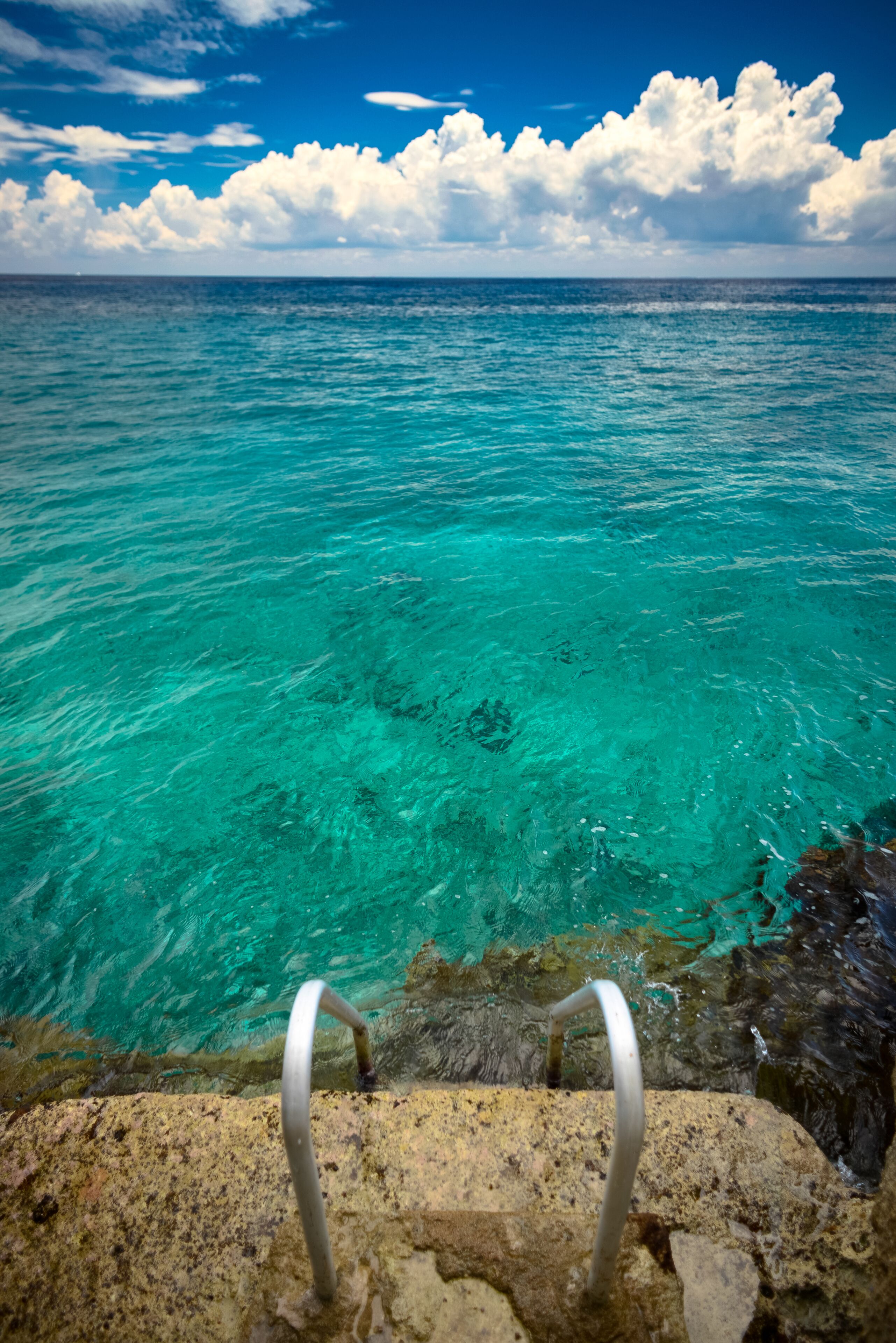 Duidelijk uitzicht op turquoise zeewater met een metalen ladder die naar de rotsen leidt.